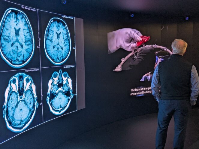 Man standing in an immersive space with a brain scan image on one portion of the screen while a video occupies another section