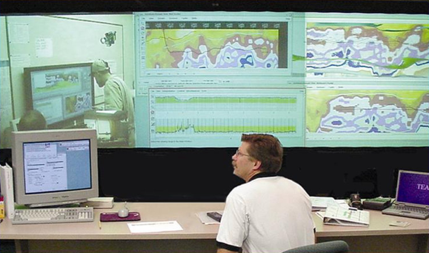 Operations center with a large wall display showing geological data, charts, and remote collaboration screens while a person works at a desk with multiple computer monitors.