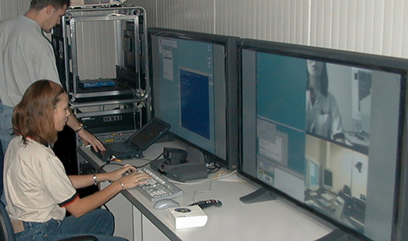 Control room with two people operating a computer workstation connected to multiple large monitors displaying video conferencing and remote monitoring screens.
