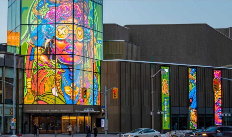 Large-scale transparent LED display integrated into the glass façade of a performing arts center, featuring a vibrant, colorful portrait that wraps around the corner of the building.
