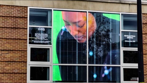 Transparent LED display installed behind a multi-story window wall, showing video artwork of a person while maintaining visibility through the glass exterior.