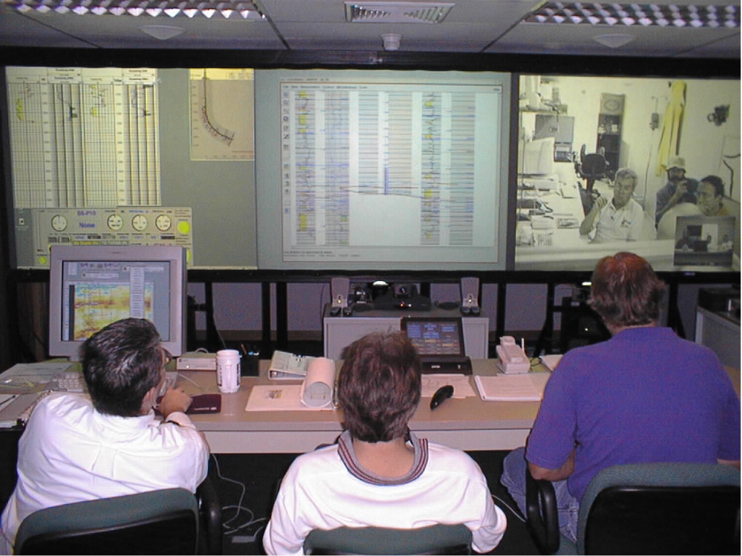 Operations control room with three people viewing a large wall display showing technical data, charts, and a remote video conference.