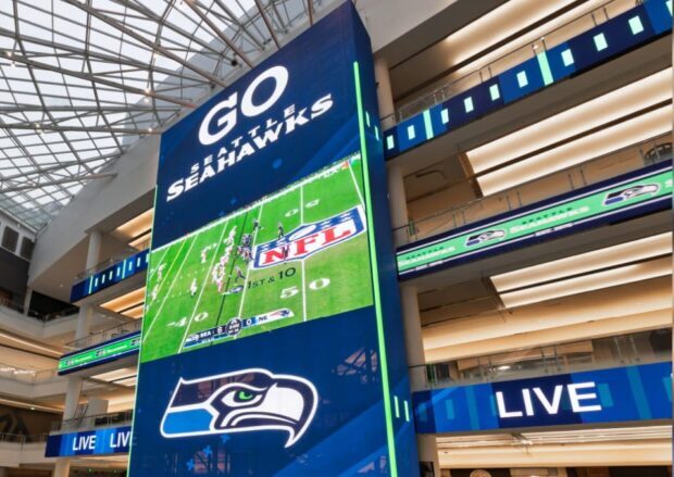 Large digital display inside a multi-level atrium showing Seattle Seahawks branding and a live NFL football broadcast, with illuminated architectural signage surrounding the screen.